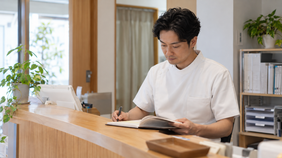 施術所の受付で経費の記録を確認する男性のAI生成イメージ